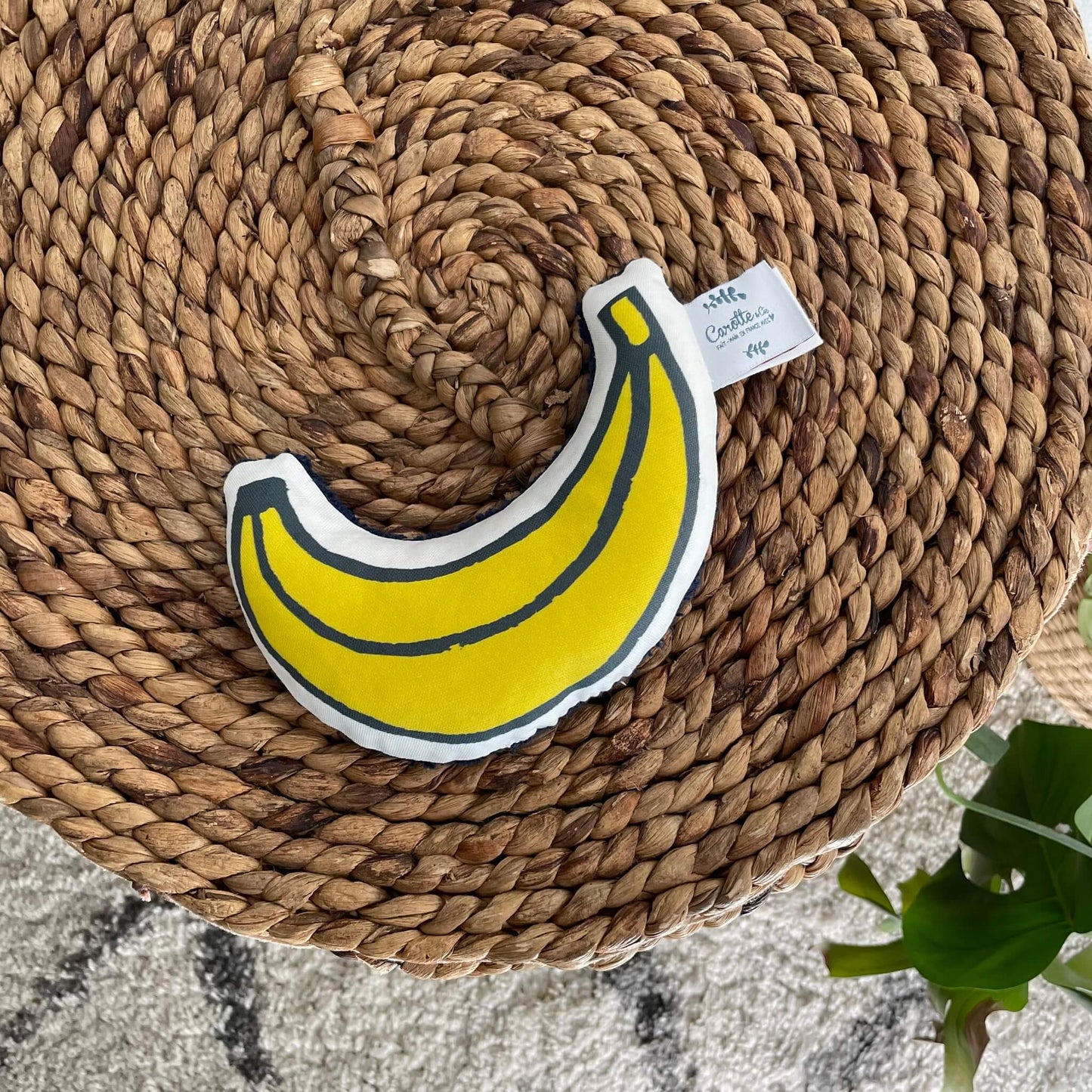 hochet banane bébé en coton bio présenté sur un pouf en rotin
